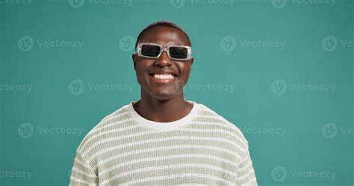 smiling person in sunglasses and jacket on street with building in the background